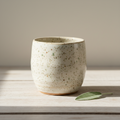 Hand-thrown speckled cream stoneware mug with sage leaf on a wooden surface, soft natural light