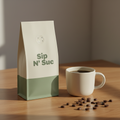 Cream paper coffee bag with sage green band and Sip N' Suc logo, ceramic mug, and scattered coffee beans on a warm wooden surface